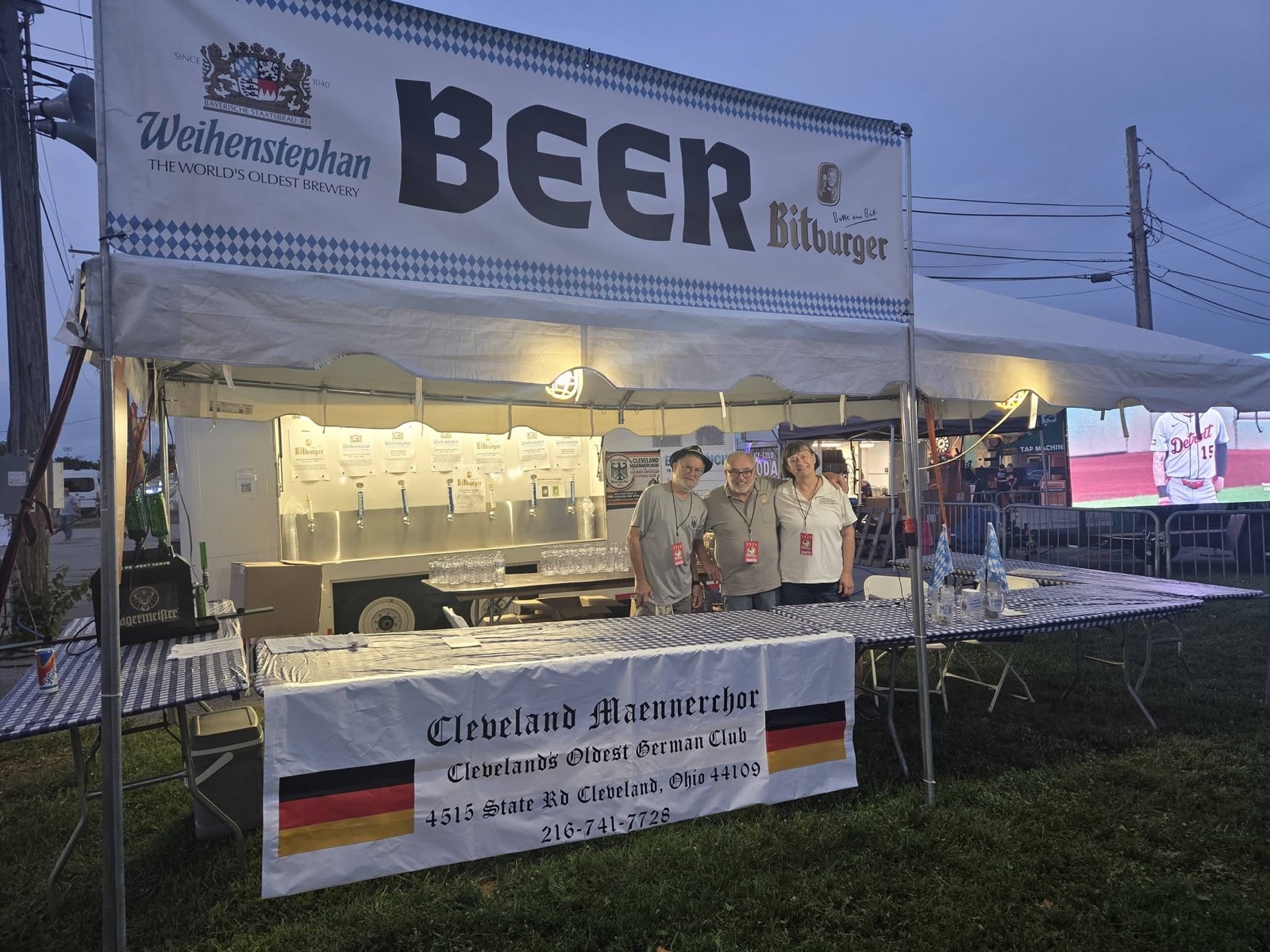 Three Cleveland Maennerchor volunteers standing behind the beer taps at dusk with the club banner on the table
